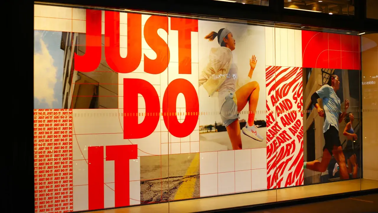 Slogan "Just Do It" in rosso su vetrina Nike, con atleti in corsa. Esempi di slogan motivazionali.