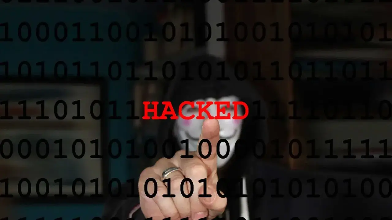 Persona con maschera anonima e codice binario sullo sfondo. La parola "HACKED" in rosso indica il significato di hackerato.