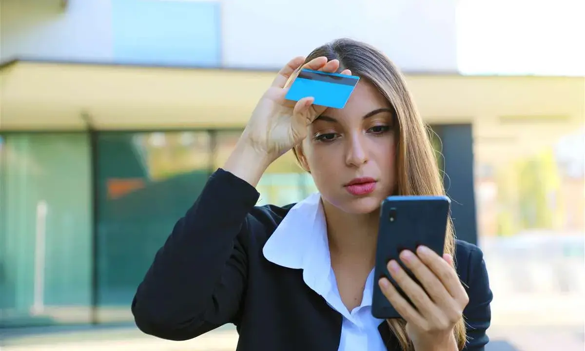 Donna preoccupata guarda il telefono con una carta in mano, temendo una carta clonata.