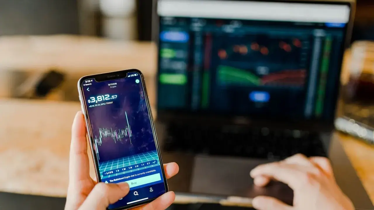 Mani che controllano grafici di Bitcoin su smartphone e laptop, un promemoria visivo delle potenziali truffe trading online.