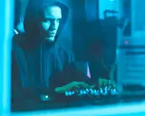 Ragazzo con cappuccio che digita su una tastiera illuminata, immerso nel blu. Un'immagine che evoca il mistero di come diventare hacker.