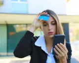 Donna preoccupata guarda il telefono con una carta in mano, temendo una carta clonata.
