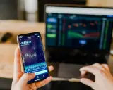 Mani che controllano grafici di Bitcoin su smartphone e laptop, un promemoria visivo delle potenziali truffe trading online.