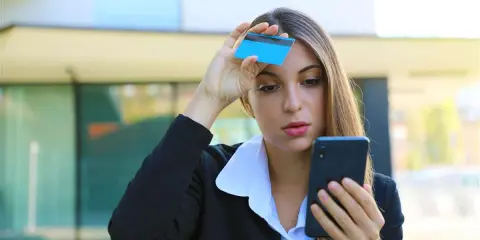 Donna preoccupata guarda il telefono con una carta in mano, temendo una carta clonata.