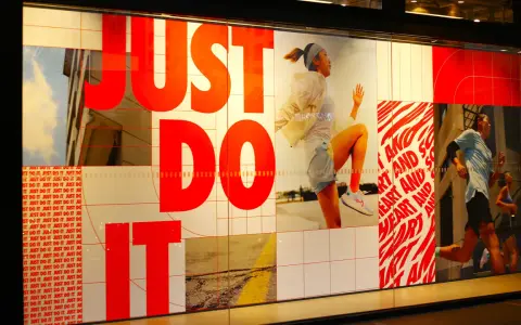 Slogan "Just Do It" in rosso su vetrina Nike, con atleti in corsa. Esempi di slogan motivazionali.