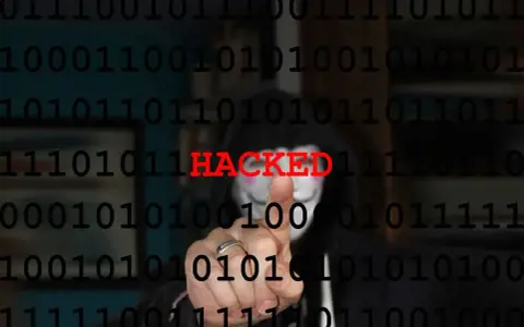 Persona con maschera anonima e codice binario sullo sfondo. La parola "HACKED" in rosso indica il significato di hackerato.
