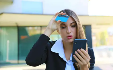 Donna preoccupata guarda il telefono con una carta in mano, temendo una carta clonata.
