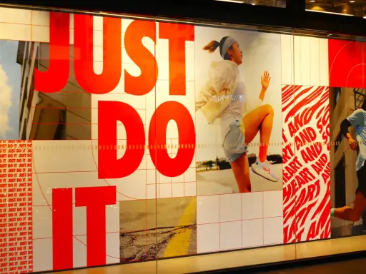 Slogan "Just Do It" in rosso su vetrina Nike, con atleti in corsa. Esempi di slogan motivazionali.