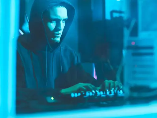 Ragazzo con cappuccio che digita su una tastiera illuminata, immerso nel blu. Un'immagine che evoca il mistero di come diventare hacker.