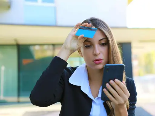 Donna preoccupata guarda il telefono con una carta in mano, temendo una carta clonata.