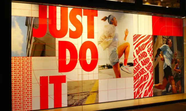 Slogan "Just Do It" in rosso su vetrina Nike, con atleti in corsa. Esempi di slogan motivazionali.
