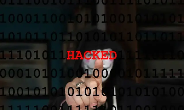Persona con maschera anonima e codice binario sullo sfondo. La parola "HACKED" in rosso indica il significato di hackerato.