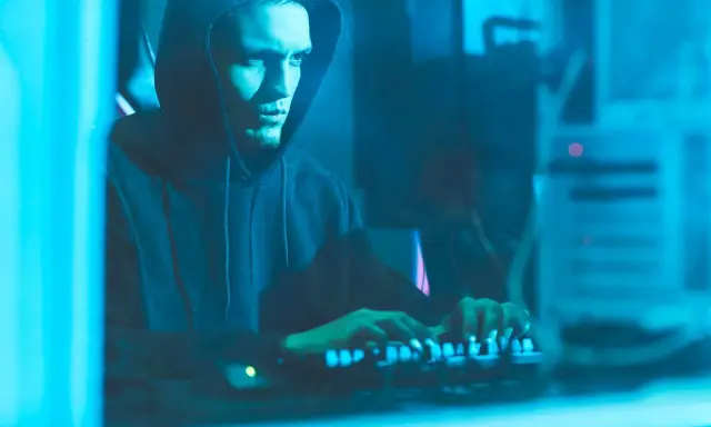 Ragazzo con cappuccio che digita su una tastiera illuminata, immerso nel blu. Un'immagine che evoca il mistero di come diventare hacker.