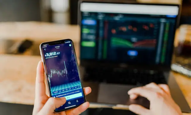 Mani che controllano grafici di Bitcoin su smartphone e laptop, un promemoria visivo delle potenziali truffe trading online.