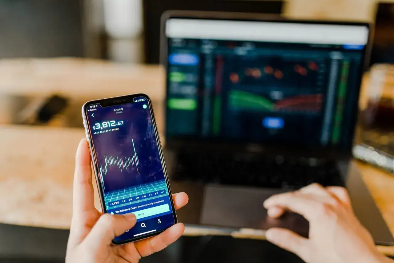Mani che controllano grafici di trading su smartphone e laptop, un promemoria visivo delle potenziali truffe trading online.