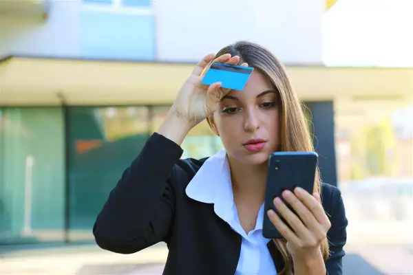 Donna preoccupata guarda il telefono con una carta in mano, temendo una carta clonata.